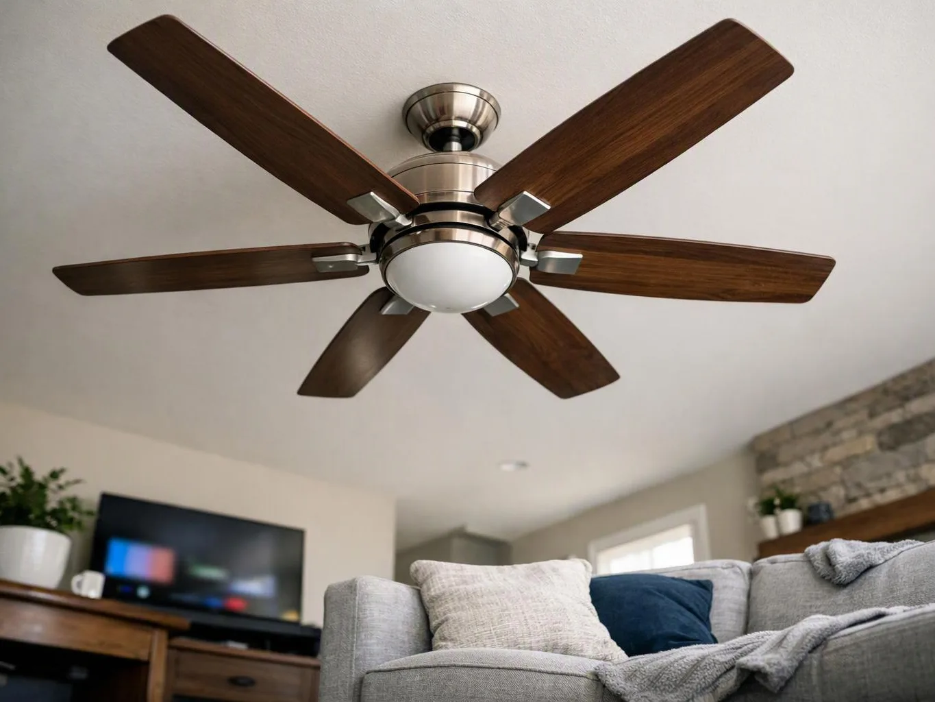 A modern ceiling fan with wooden blades hangs above a cozy living room with a sofa and TV