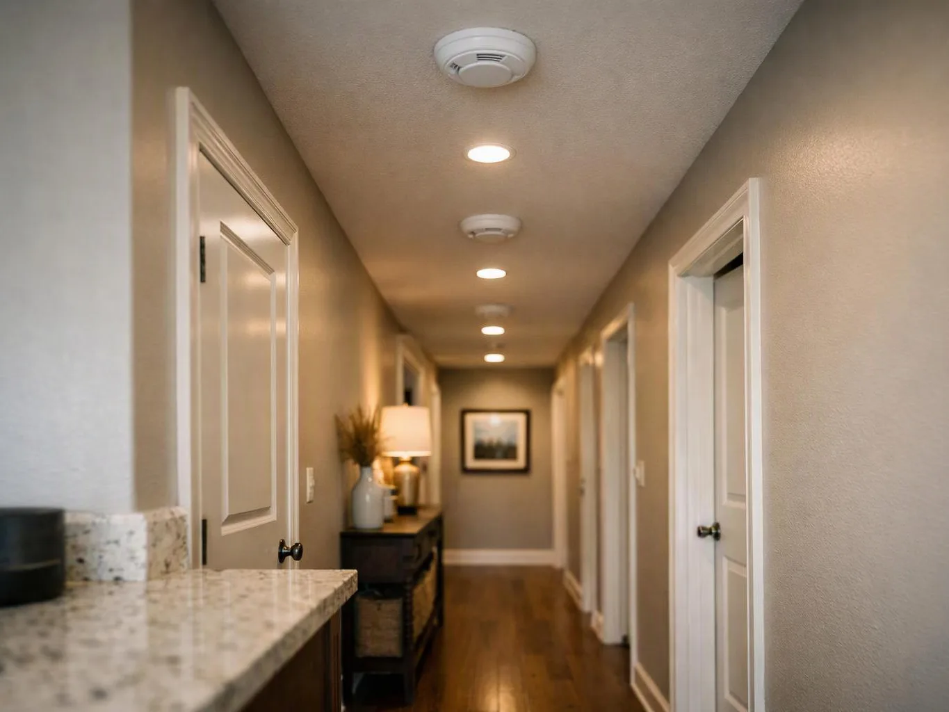 A narrow hallway with white doors, recessed lights, and a small console table leads toward a framed picture at the end